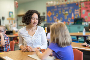 A young girl looks up from her desk to talk with her teacher, who has come over to check on how she's doing with her writing assignment. The shot is focused on the teacher and is over the left shoulder of the student.